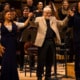 Three people holding hands, taking a bow, in front of an orchestra on stage.