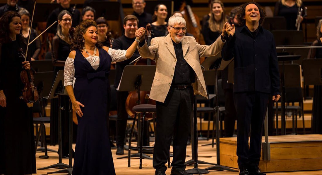 Three people holding hands, taking a bow, in front of an orchestra on stage.
