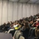 A person holding a microphone, speaking to a lecture hall full of people.