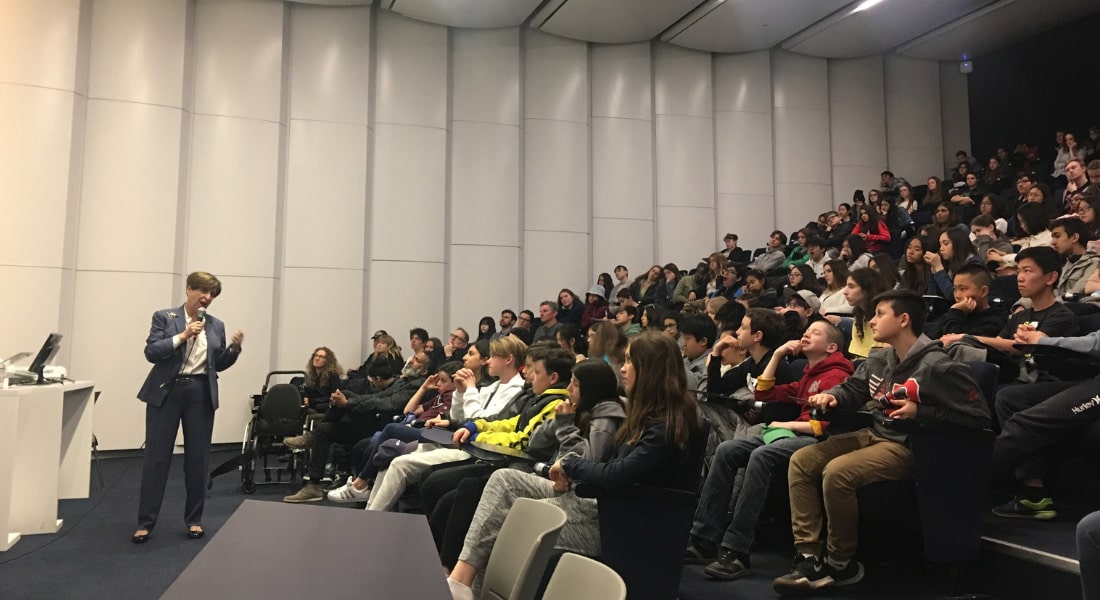 A person holding a microphone, speaking to a lecture hall full of people.