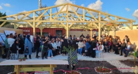 Large group of people, seated and standing in front of a partially built outdoor structure, listening someone giving a speech.
