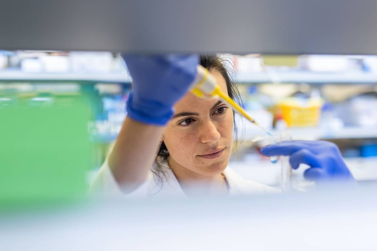 A person with blue rubber gloves and a lab coat dropping liquid into a test tube.