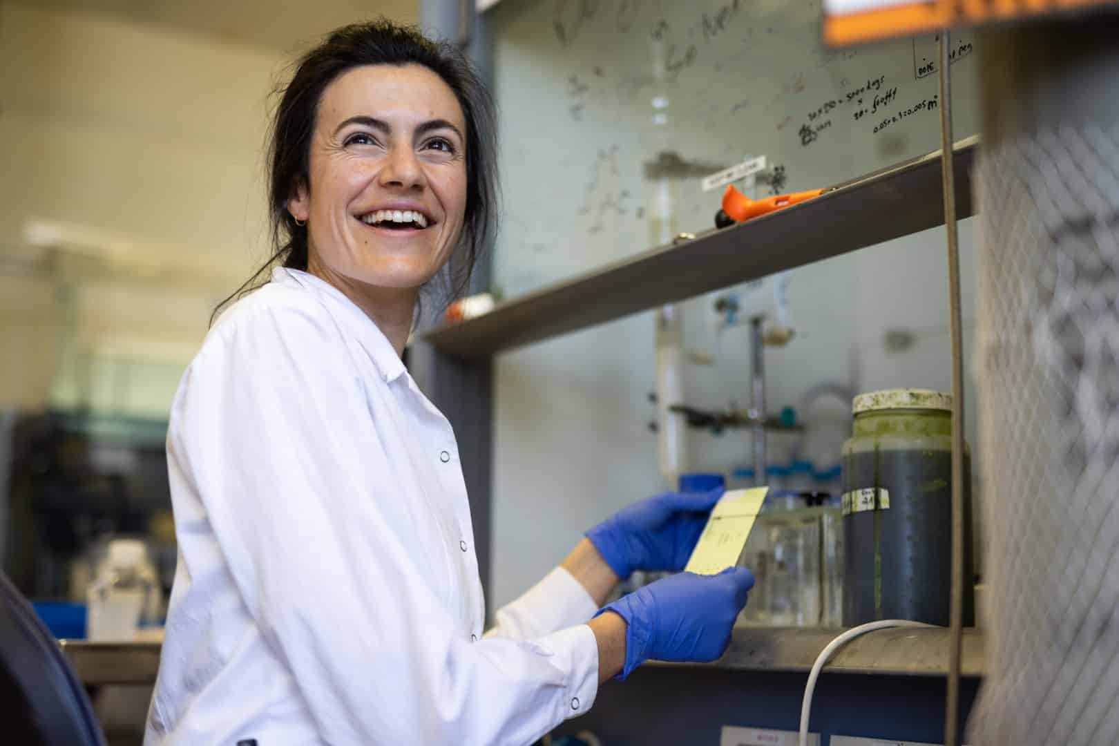 A person with blue gloves and a lab coat smiling while holding a yellow card.